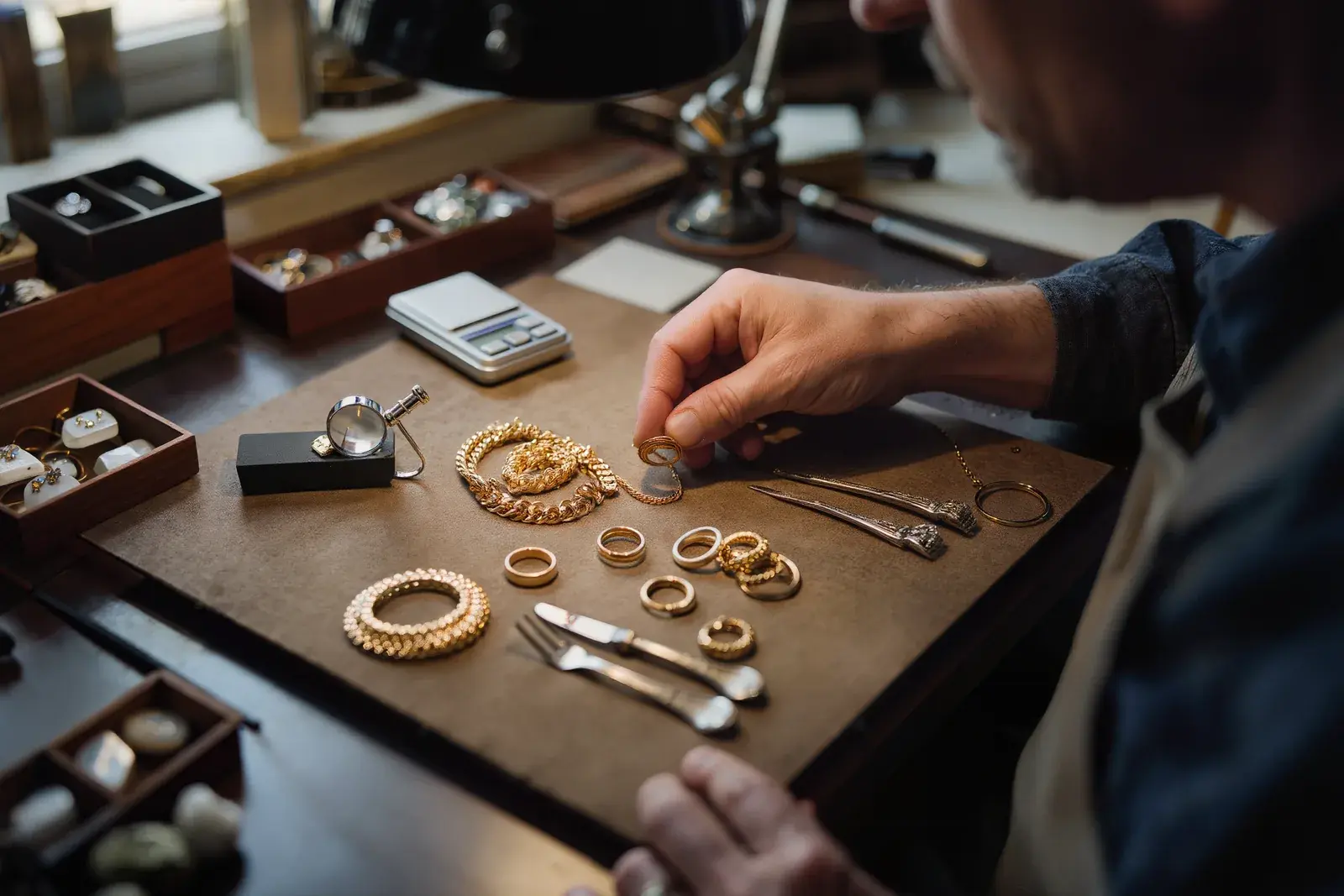 Gold and silver pieces sorted into groups on a desk mat beside a jeweller's loupe and a scale